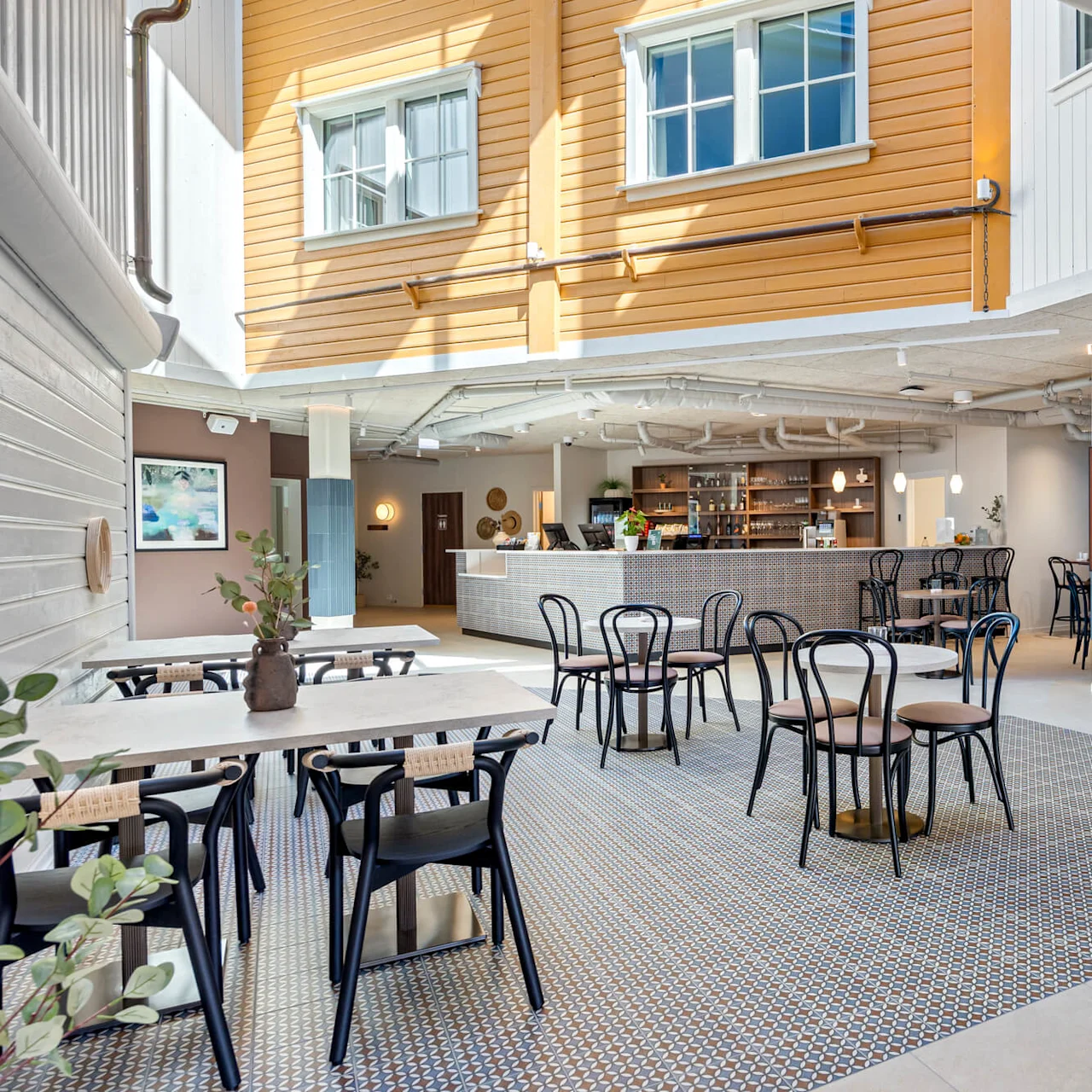 A well-lit spacious cafe interior with wooden tables, metal chairs, a counter in the back, and potted plants, inside a building with wooden siding and large windows.