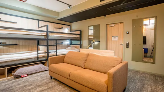 A brown sofa sits in a modern room with two sets of bunk beds against the wall, reflected in two mirrors. A door and lamp are nearby. No text present.