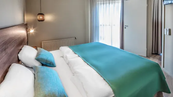 A neatly made bed with turquoise bedding and cushions sits in a tidy modern bedroom. A hanging lamp illuminates the scene, and sheer curtains cover a sunlit window. 