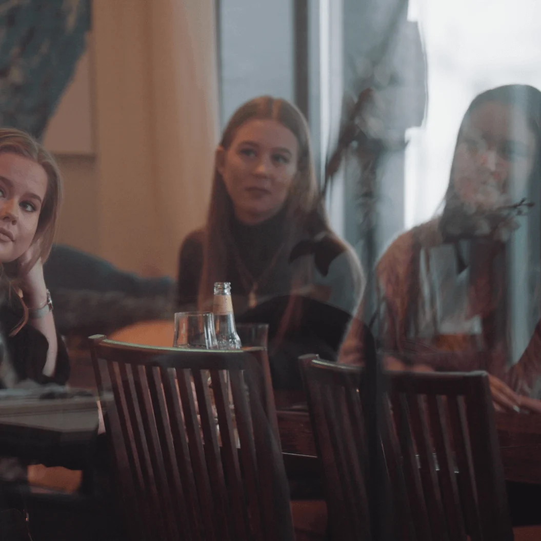 En kvinne sitter på en kafé og hviler haken i hendene, ser ettertenksom ut, med en annen kvinne i bakgrunnen.