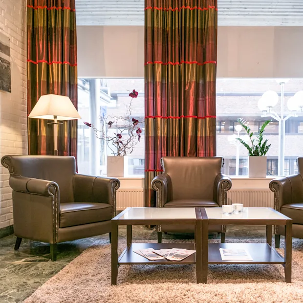 Three brown leather armchairs surround a coffee table holding magazines in a cozy lounge. A lamp stands beside, with large windows and vibrant curtains providing natural light.