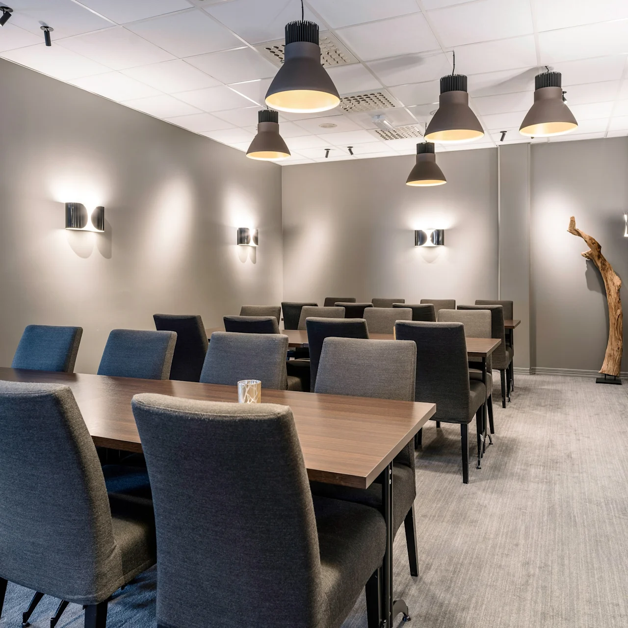 Tables and chairs are arranged neatly in a conference room with gray walls and soft lighting. Decorative sconces and a wooden sculpture add a touch of elegance.