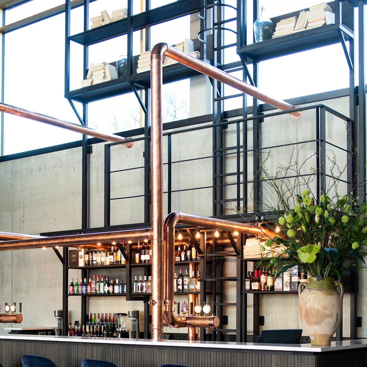 A modern bar with four dark blue high chairs, featuring copper piping and shelves of various bottles, situated in a spacious, industrial setting with a large green plant.