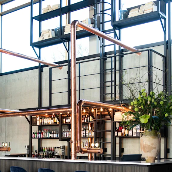 En modern bar med fyra mörkblå höga stolar, kopparrör och hyllor med olika flaskor, belägen i en rymlig, industriell miljö med en stor grön växt.