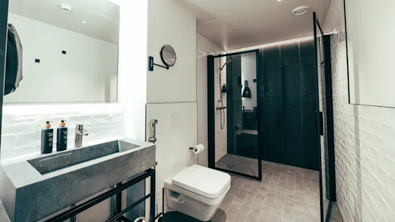 Et moderne baderom har en grå steinvask med to flasker, en vegghengt toalett og en glassdør til dusjen. Hvite flislagte vegger forsterker det elegante designet.