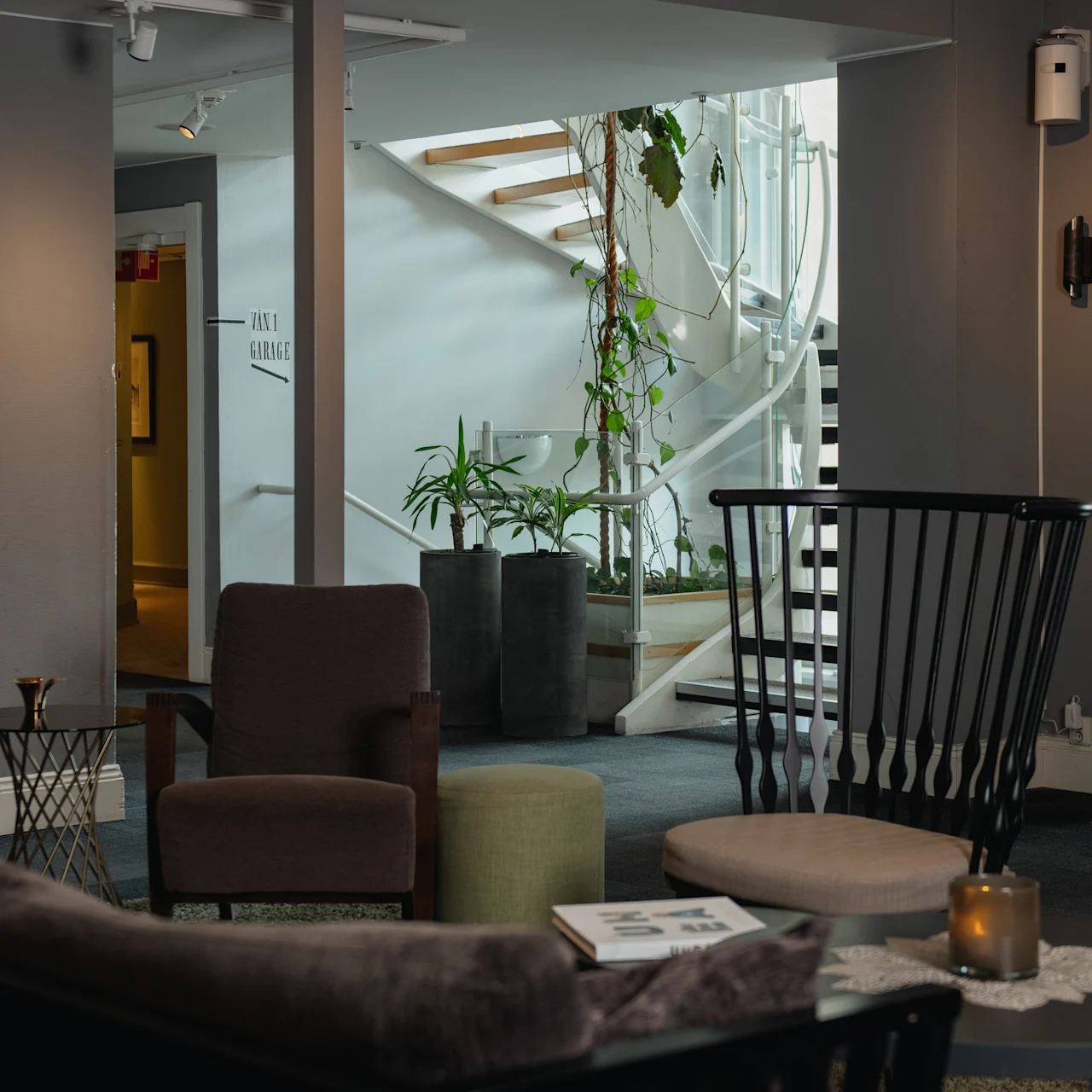 Comfortable seating area with armchairs and small tables; candle on one table. Staircase with potted plants in the background. Walls in gray tones. Text on wall: “JOLY ORANGE”.