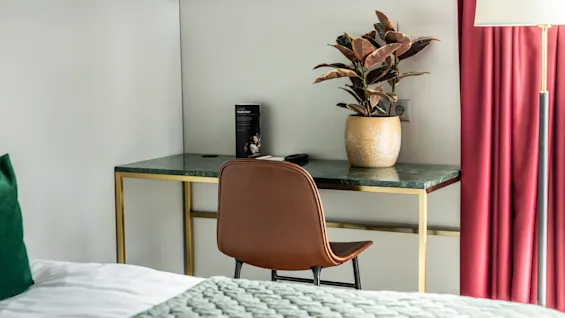Ett mysigt hörn i ett hotellrum med ett elegant skrivbord, en krukväxt, en broschyr med titeln "Your Stay" och en brun stol. Den varma känslan förstärks av röda gardiner och ett mjukt grönt överkast.