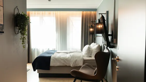 Et koselig hotellrom med en komfortabel seng, en stilig stol og en grønn plante, badet i naturlig lys fra vinduet.