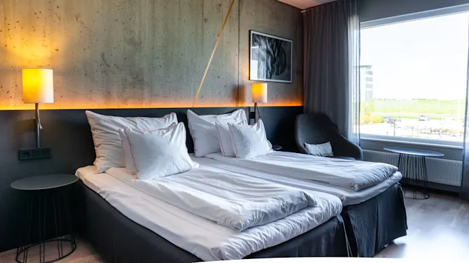 En pent oppredd seng med hvite sengetøy og puter ved siden av en stilfull lampe, plassert i et moderne, godt belyst hotellrom med store vinduer og utsikt utenfor.