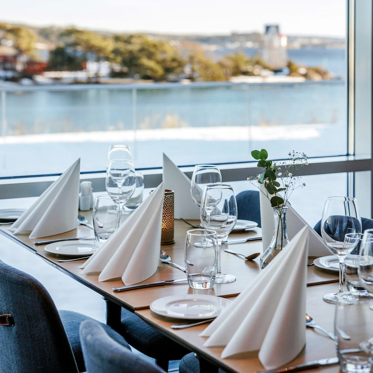 A dining table is set with folded napkins, glassware, and cutlery, overlooking a snowy landscape and serene body of water through large windows.  