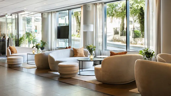 A modern lounge area with plush white sofas arranged around small tables, set in a sunlit room with large windows overlooking greenery. No text is visible.