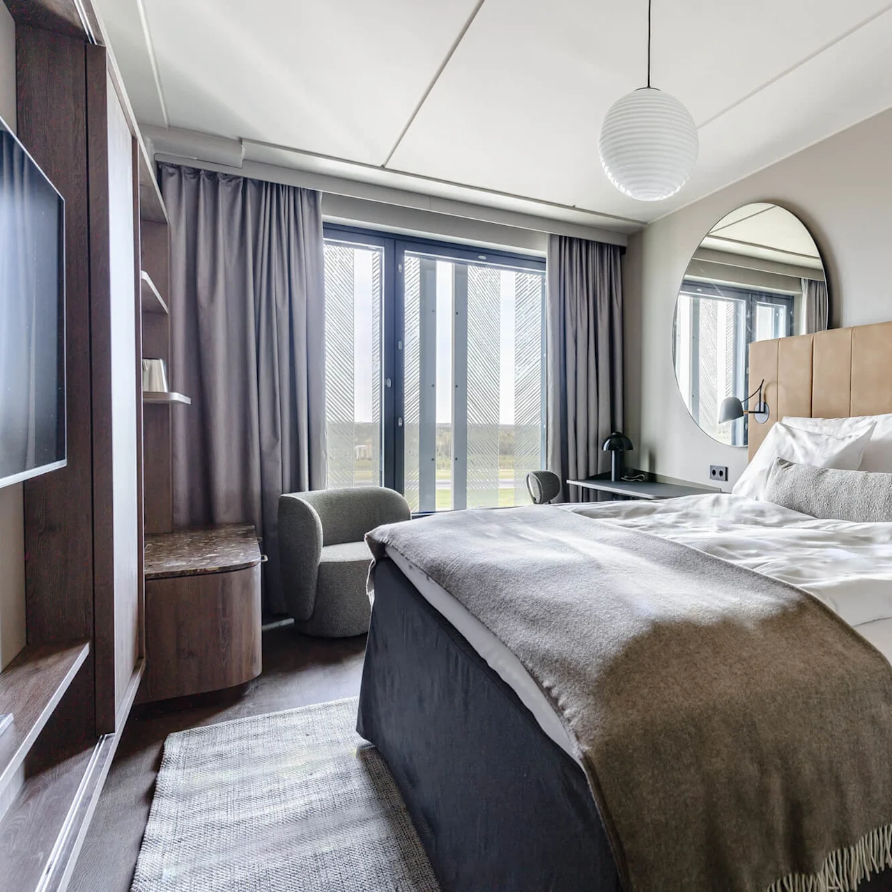 Bed neatly made with white linens and gray blanket, positioned in a modern bedroom with brown leather headboard, large circular mirror, and window covered by gray curtains.