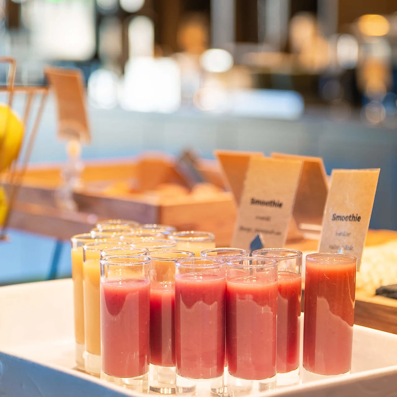 Glas med smoothies står i en hvid bakke; gule og pink varianter. Sløret baggrund antyder en café. Synlig tekst: "Smoothie", "met", "bleuet", "Smoothie", "nectar", "multivitam!"