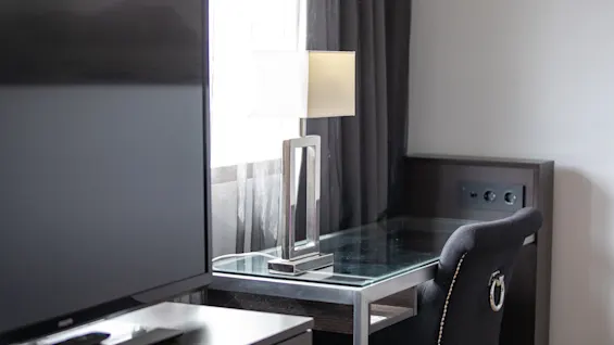 En stor flatskjerm-TV står på et trebord ved siden av et glassbord med en kantet bordlampe. En myk stol vender mot bordet. Mørke gardiner rammer inn et vindu.