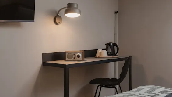 Viihtyisä hotellihuoneen työpöytäalue lampun, radion, vedenkeittimen ja kuppien kera, valmiina rentouttavaan oleskeluun.