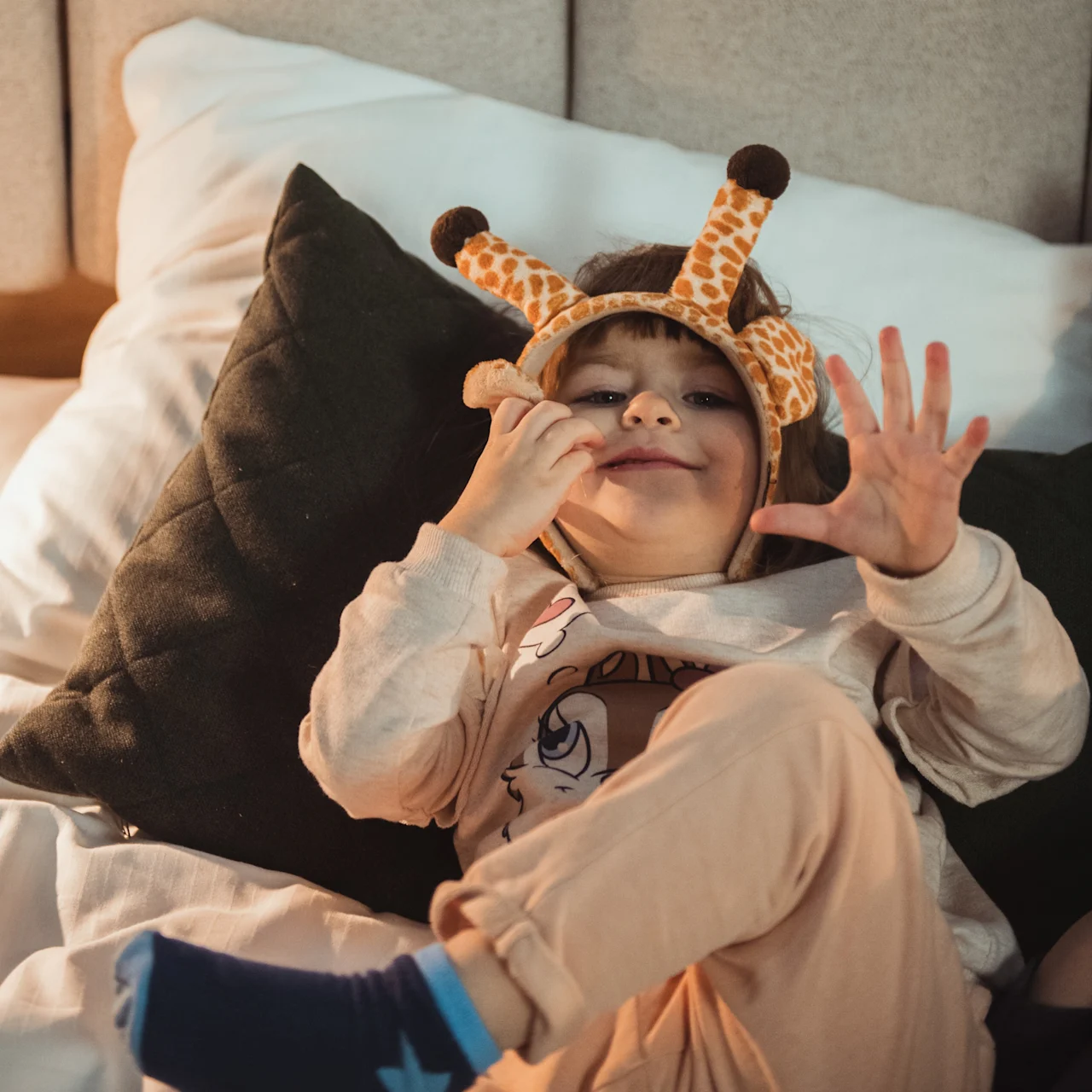 Et barn med et sjiraffpannebånd ligger lekent på en seng, smiler og holder opp en hånd.