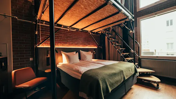 A modern hotel room with a bed, a loft area above, and a staircase leading up. A large window brightens the industrial-style space.