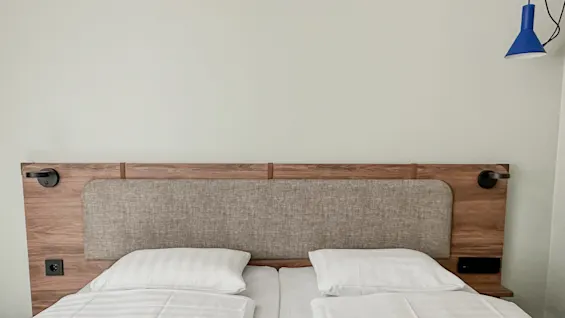 En seng på et hotellrom er pent oppredd med hvite laken og to puter, har en trehodegavl og tilhørende lamper, i et minimalistisk rom med lyse vegger.