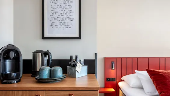 A hotel room's side table with a coffee maker and accessories against a wood-panel headboard.
