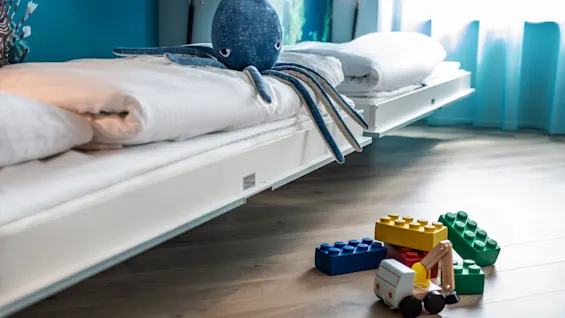 A stuffed octopus sits on a white bed in a blue-themed room; toy blocks and a toy truck are scattered on the wooden floor.