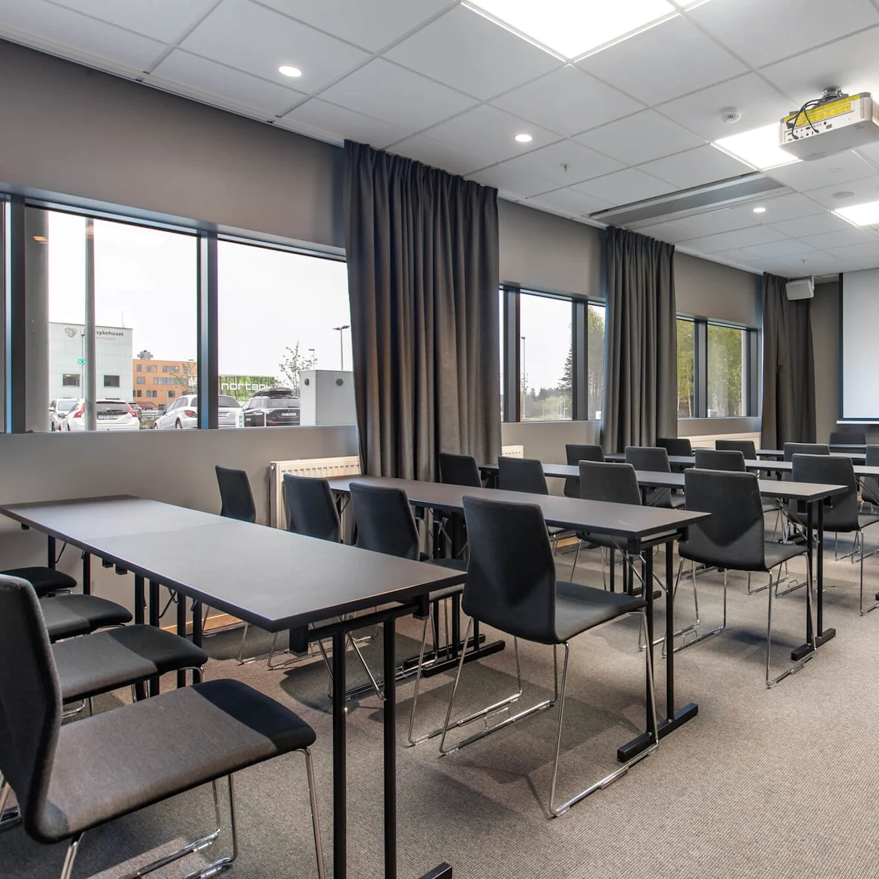 Conference room with rows of empty black chairs facing a large white projection screen. The room has gray curtains, windows on the left, and bright overhead lights.