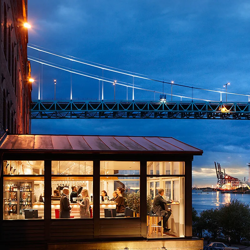 Restaurant patrons dine inside a glass-walled building beside a river. A bridge spans overhead, while a large ferry labeled "Stena Line" passes by in the evening light.