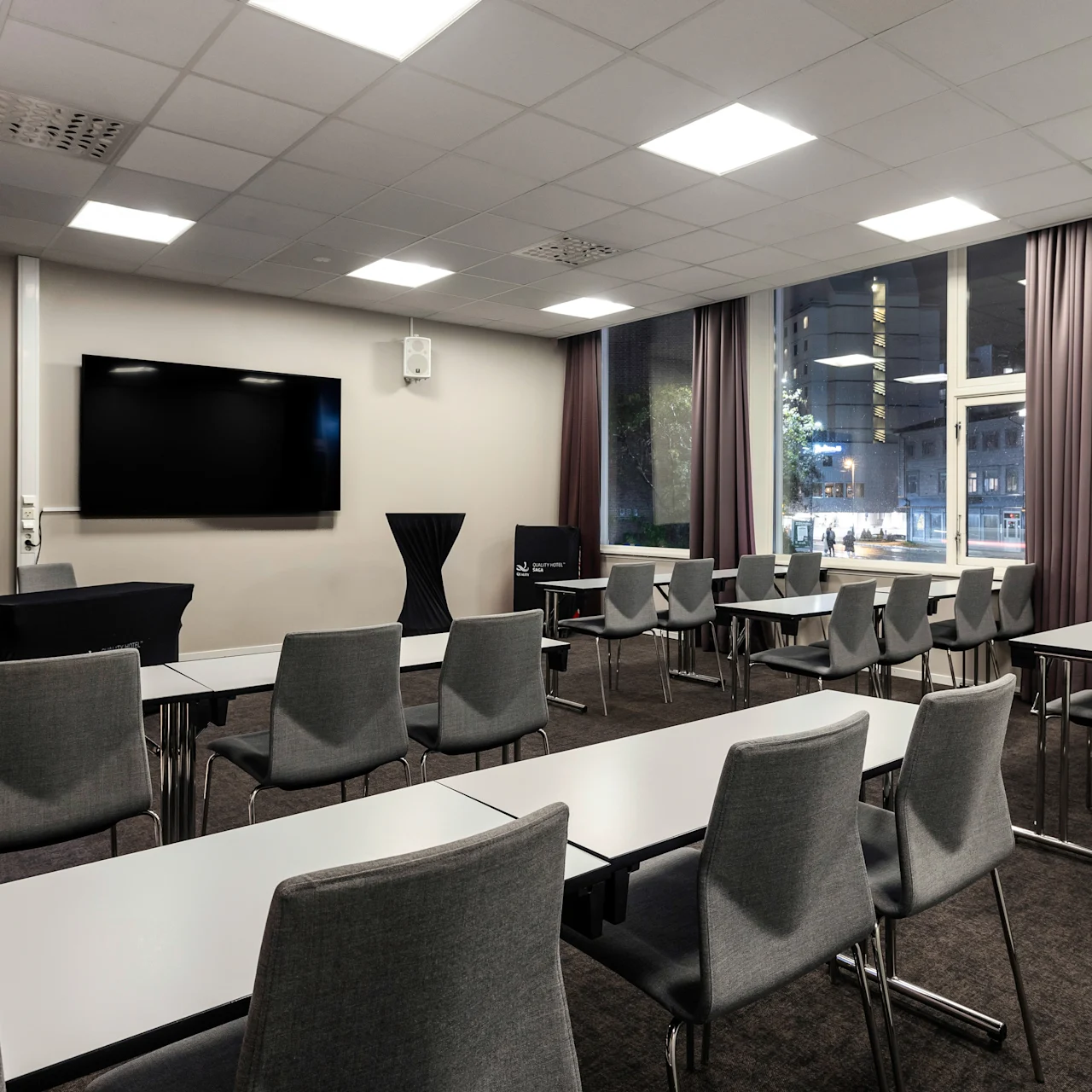Modern conference room with neatly arranged gray chairs facing a wall-mounted screen. Tall windows overlook a cityscape. Text: “QUALITY HOTEL SAGA Tromsø.” 