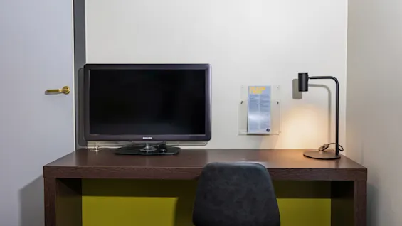 TV on a desk; Philips, turned off. A document is clipped to the wall beside a lit lamp. A chair faces the desk. The room has white walls.