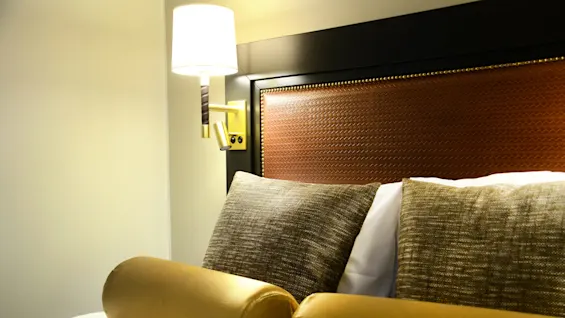 A neatly made bed with a gold bolster, a lamp above in a well-lit room.