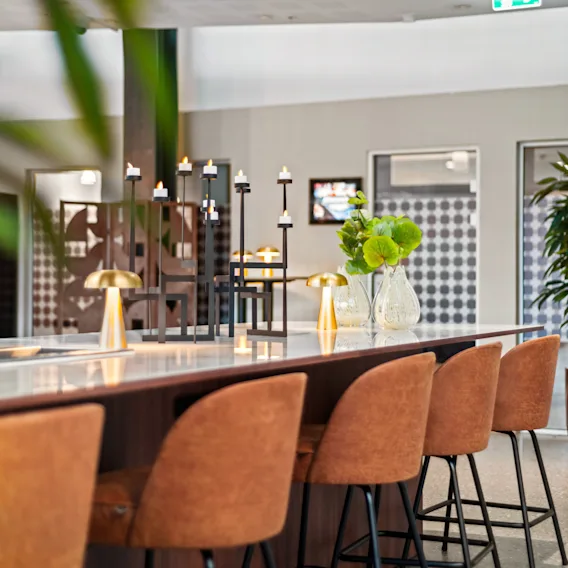 En lang, hvit benkeplate med brune stoler er pyntet med gullfargede lamper og vaser med grønne planter. Den står i et moderne, åpent rom med dekorative planter og hyller.
