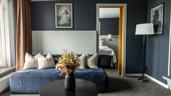 Ett mysigt vardagsrum med en elegant soffa dekorerad med mönstrade kuddar. Ett stort fönster fyller rummet med ljus. Ett bord håller en vas med blommor. Sovrummet syns genom en öppen dörr med en mjuk säng.