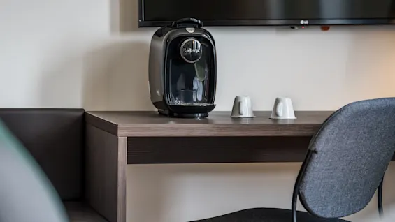 A black coffee machine labeled "Löfbergs" sits on a wooden table next to two inverted mugs. A wall-mounted TV is above, with a chair positioned nearby.