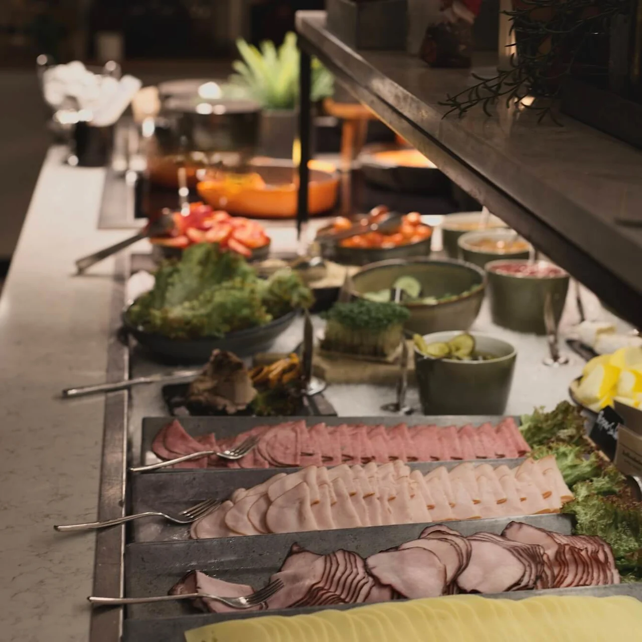 A buffet with a variety of cold cuts and cheese slices neatly arranged, tongs ready for use, in a dimly lit setting.