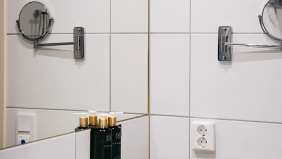 Väggmonterade runda speglar sträcker sig ovanför ett vitt handfat. Tre toalettartiklar märkta "hand & body" står på handfatet. Ett dubbeluttag syns i kakelväggen. 