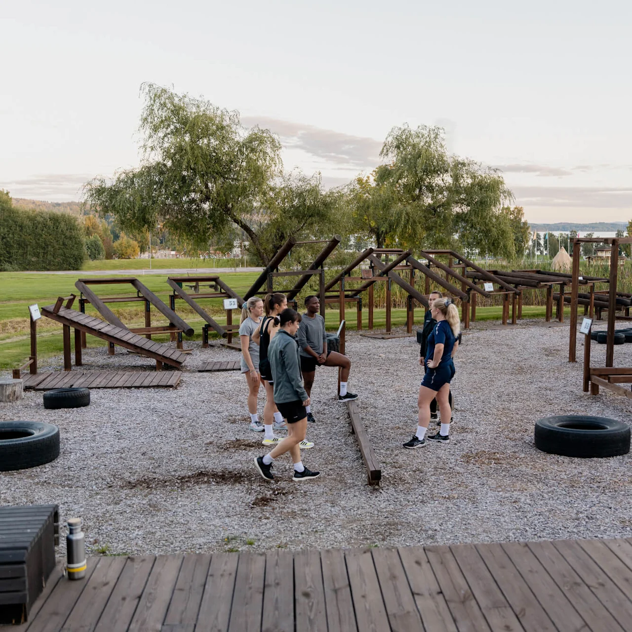 En gruppe mennesker står på en treplattform i en utendørs treningspark, omgitt av treningsapparater og trær med et frodig landskap. Store dekk er spredt rundt.