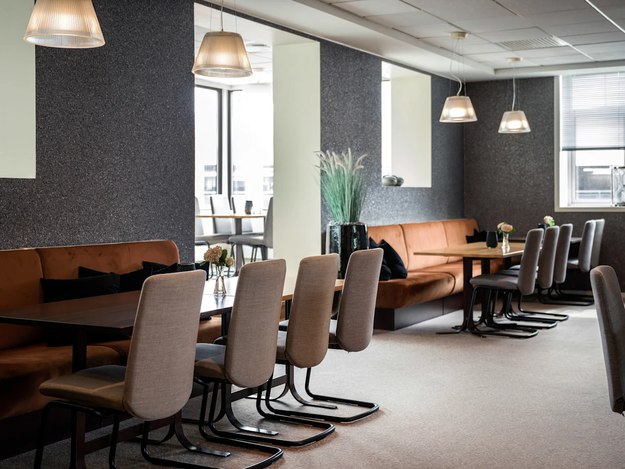 A modern dining area with comfortable seating, including banquettes and chairs around wooden tables, under soft pendant lighting.