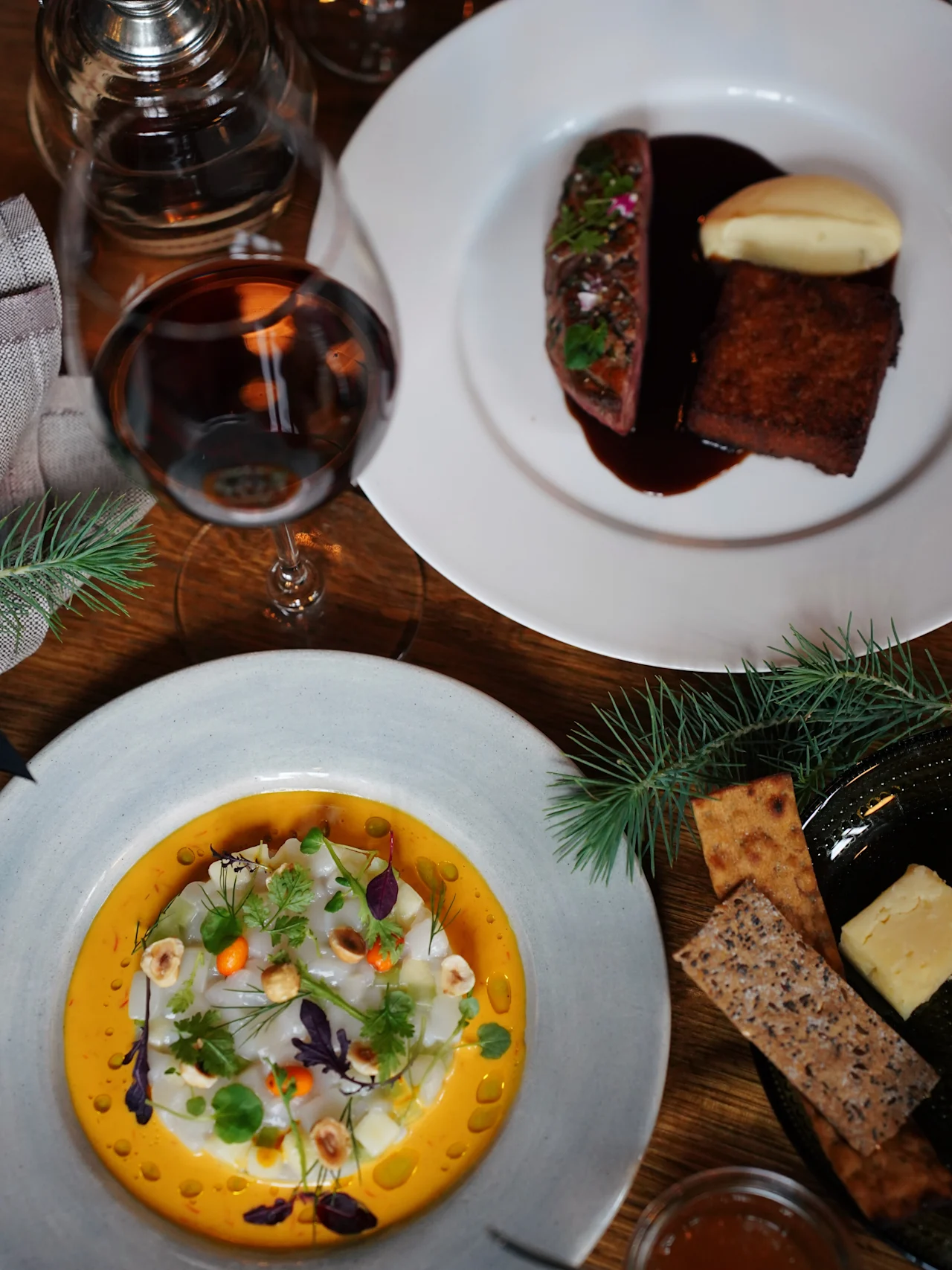 A table with gourmet dishes, including a steak with sauce and purée, a plate of colorful appetizer with greens, and wine glasses. Fir branches decorate the setting.