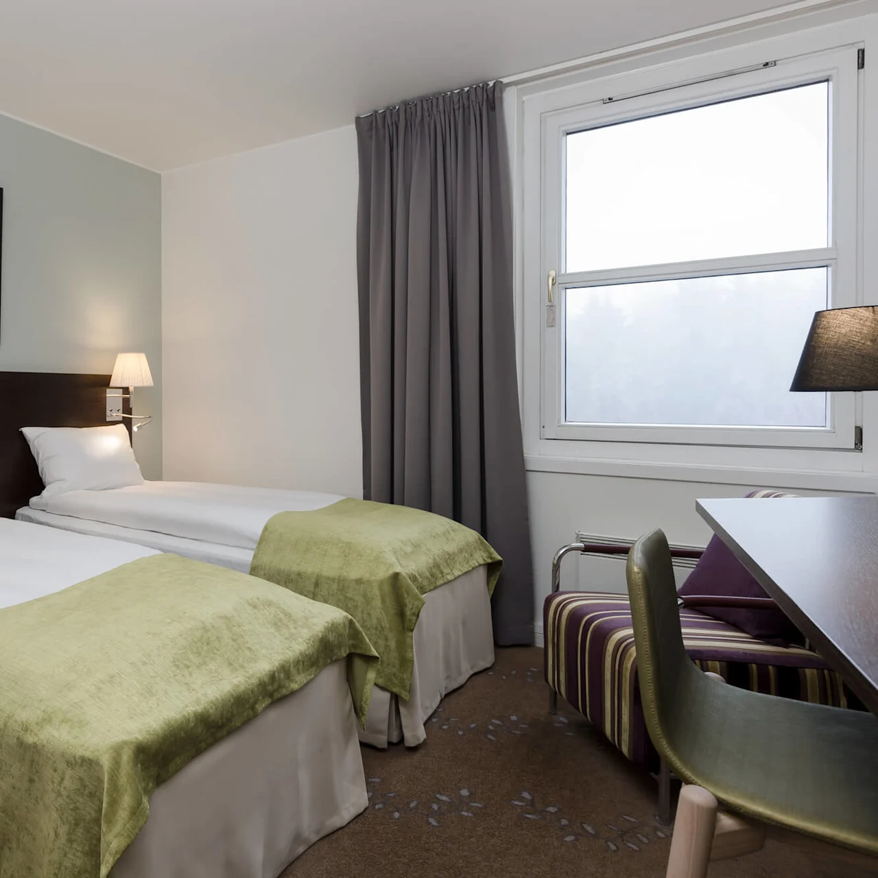 Two neatly made twin beds with green throws are beneath wall lamps in a softly lit hotel room with a window, curtains, and a desk with a chair.