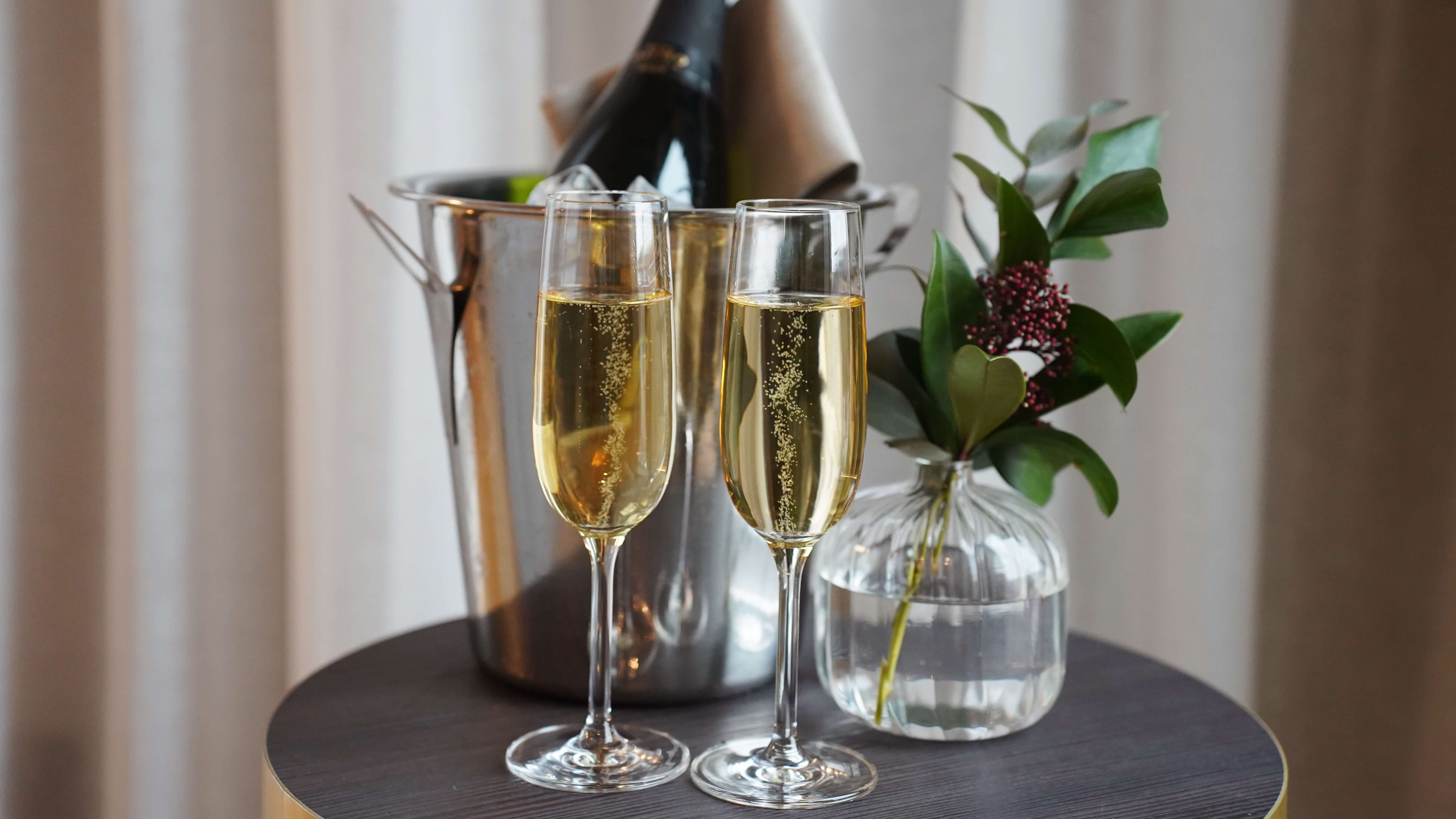 Champagne i glass med en flaske i en isbøtte og en vase med grønt, plassert på et trebord mot draperte gardiner.
