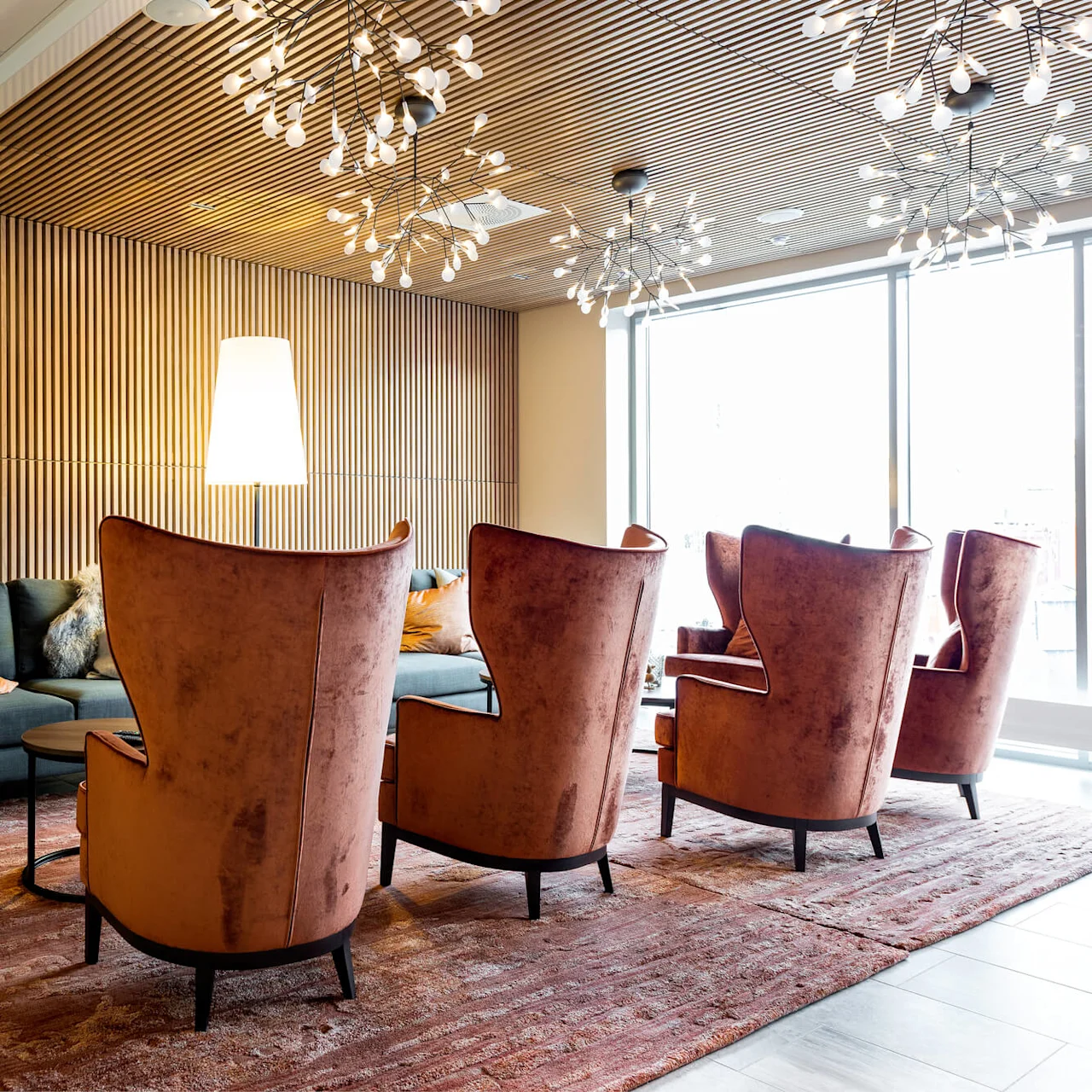 Modernit kalusteet lounge-alueella, korkeaselkäiset tuolit ja sohva, taiteelliset valaisimet yläpuolella, raidalliset puupaneelit seinillä ja isot ikkunat.