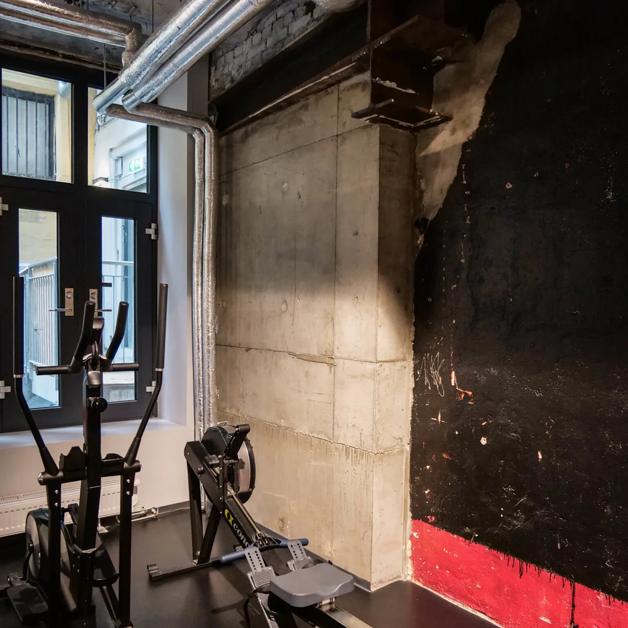 En roddmaskin och träningscykel står stilla i ett svagt upplyst gym med industristil, med synliga rör och en rå betongvägg med sektioner i rött och svart.