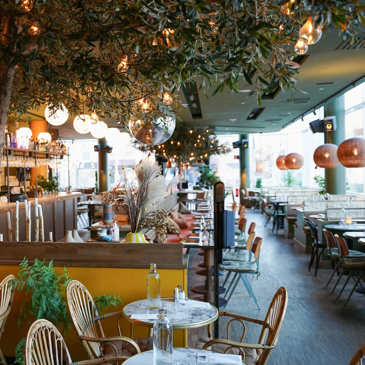 Et indendørs spiseområde med bistrostole, marmorborde, hængende lys og et træ. Stemningen er hyggelig og indbydende.