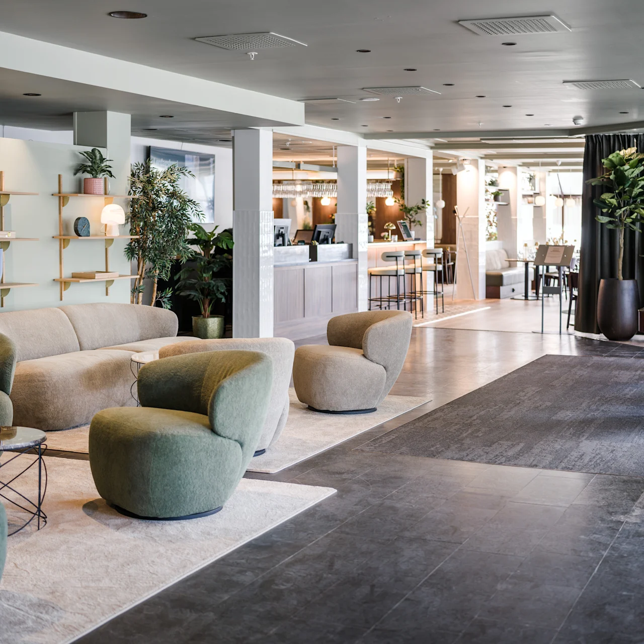 Comfortable lounge seating features plush green chairs arranged around low tables; nearby, a modern bar counter is set in a spacious, stylishly decorated area with plants and shelves.