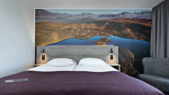 En prydligt bäddad dubbelsäng står mot en sänggavel med en naturtrogen landskapstapet. Mjuk belysning och ett mörklila överkast fullbordar den mysiga hotellrumsatmosfären.
