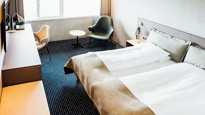 Et ryddig hotellrom har en pent oppredd seng, to lenestoler ved et rundt bord og et vindu med utsikt over en lys scene. Minimalistisk innredning med dempet belysning.