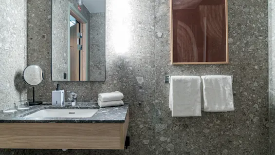 A modern bathroom with a sink, mirror, and fresh white towels, featuring grey speckled walls and a warm wooden vanity.