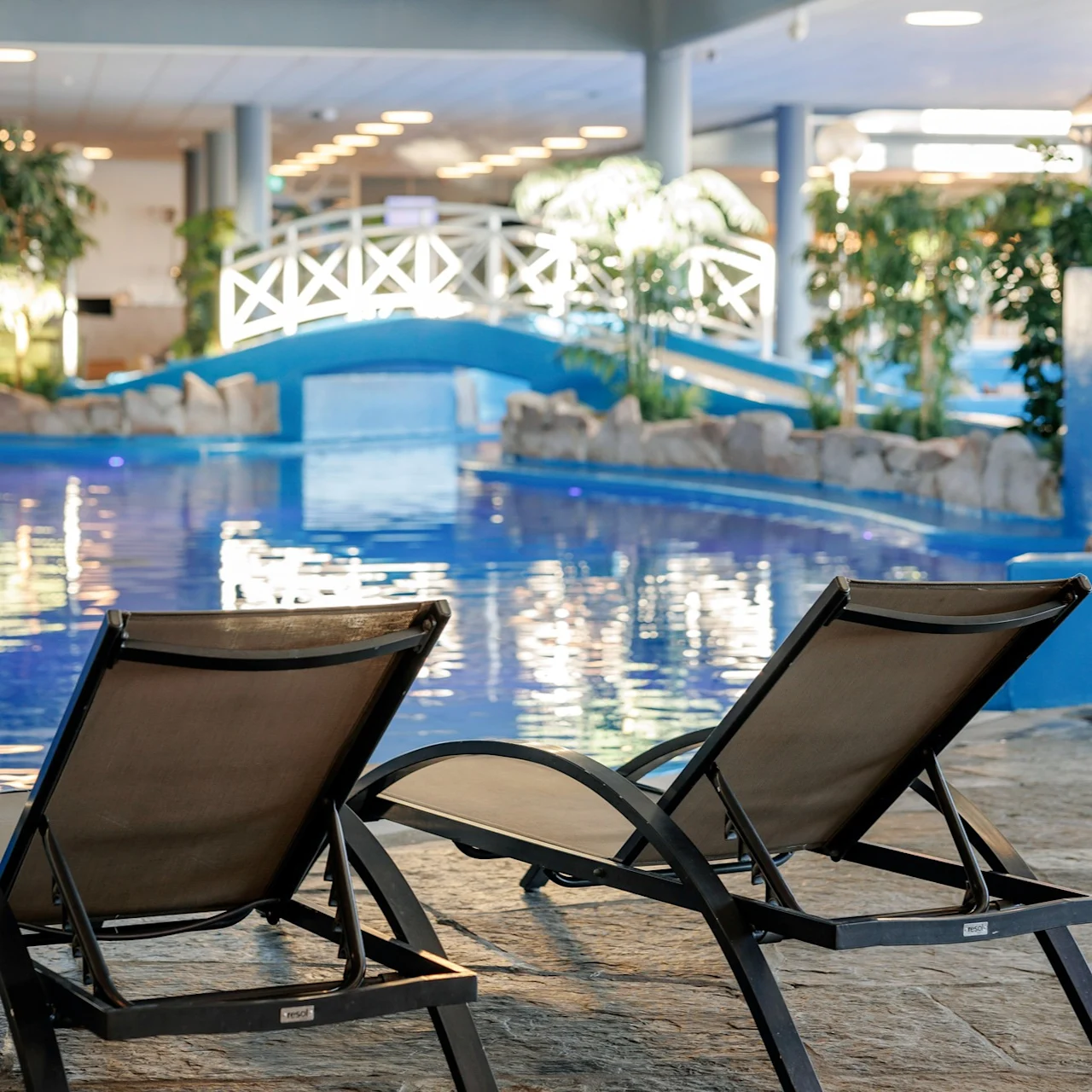 Sun loungers sit by an indoor pool, with a blue bridge and lush plants. Warm lighting creates a relaxing atmosphere.