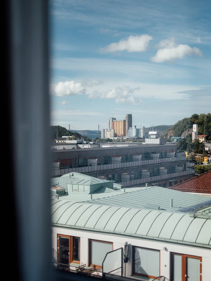 Buildings with green rooftops are viewed through a window, with a distant industrial skyline and hills under a partly cloudy blue sky.