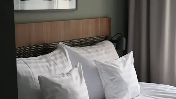A neatly made bed with white pillows and sheets, beneath a wooden headboard. Above, an abstract artwork is framed. Curtains hang beside, giving a calm, modern bedroom atmosphere.