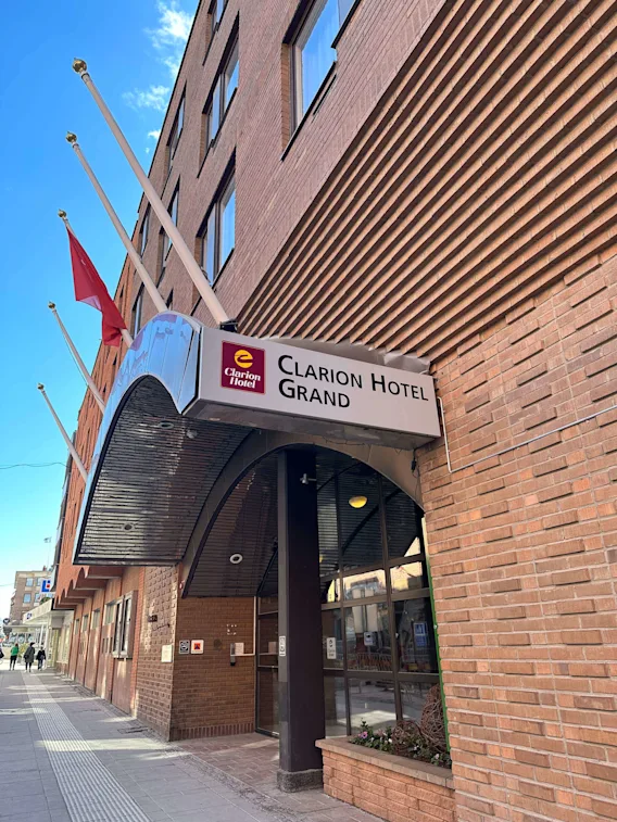 En hotellentré med en skylt som lyder "Clarion Hotel Grand." Flera flaggstänger reser sig från byggnadens tegelfasad i en stadsmiljö.